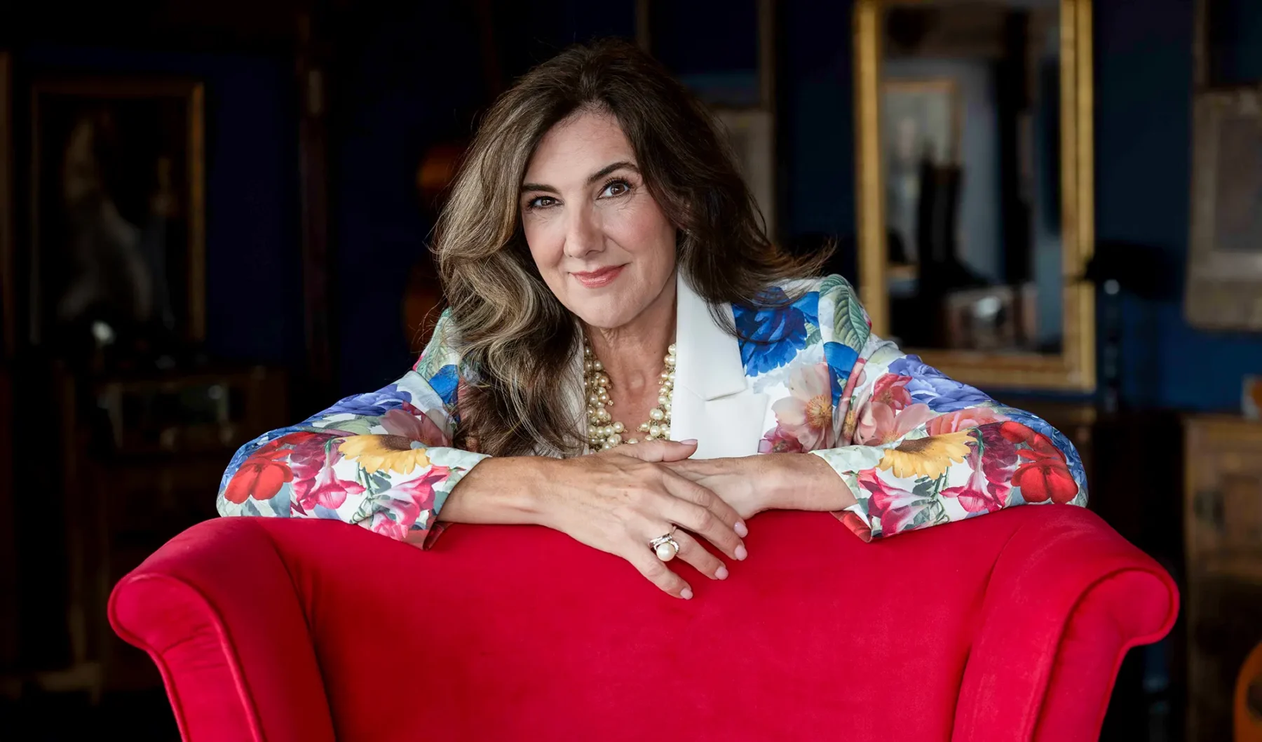 A woman with wavy brown hair, wearing a floral blazer, pearl necklace, and ring, smiles while leaning on the back of a bright red chair. The background features dark walls and framed pictures.